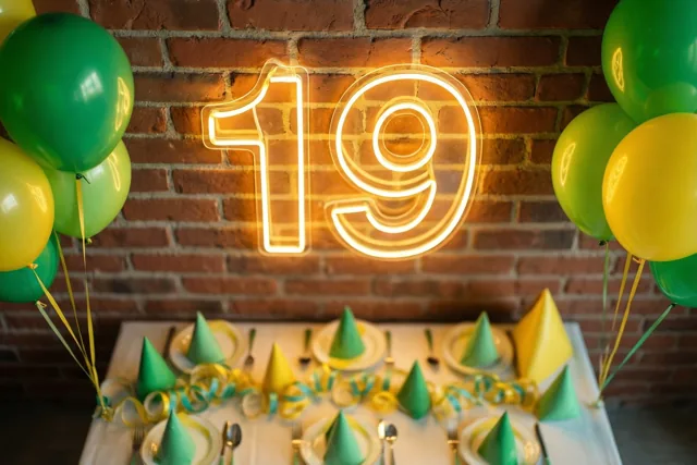 Een verlicht neongetal 19 hangt aan een rode bakstenen muur boven een feesttafel met groene en gele versiering, ballonnen en slingers.