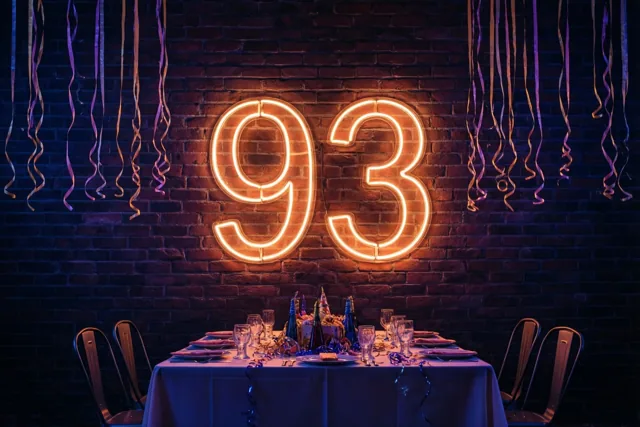 Oranje neoncijfers vormen 93 boven een feestelijk gedekte tafel tegen een donkere bakstenen muur, met paarse en gouden slingers en blauwpaars licht.