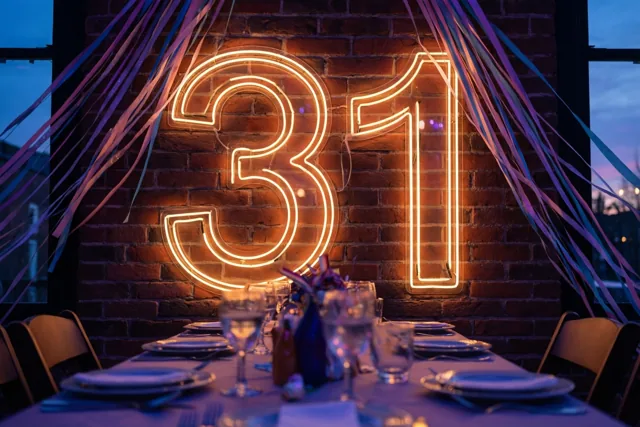 Het getal 31 licht op in neon tegen een bakstenen muur boven een gedekte feesttafel met paarse en blauwe slingers.