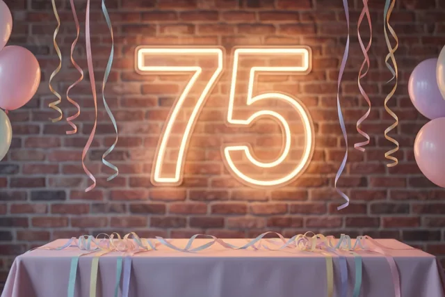 Een lichtgevende 75 in neon hangt aan een bakstenen muur boven een feesttafel met pastelballonnen en slingers.