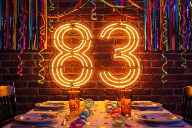 Een neonbord met 83 gloeit warm tegen een bakstenen muur boven een feesttafel met kleurrijke slingers en confetti.