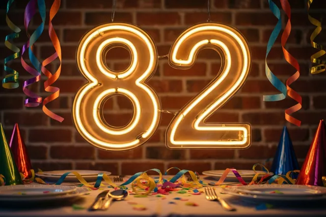 Een gloeiend neongetal 82 hangt boven een feesttafel tegen een bakstenen muur, met kleurrijke slingers en feesthoedjes.