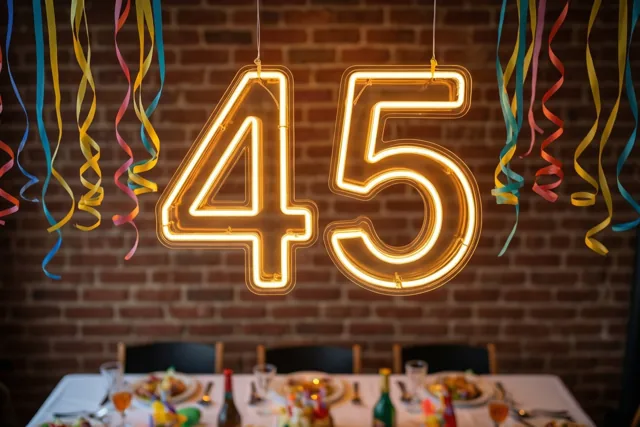 Een oranje gloeiend neongetal 45 hangt boven een feesttafel tegen een bakstenen muur, terwijl kleurrijke serpentines naar beneden vallen.