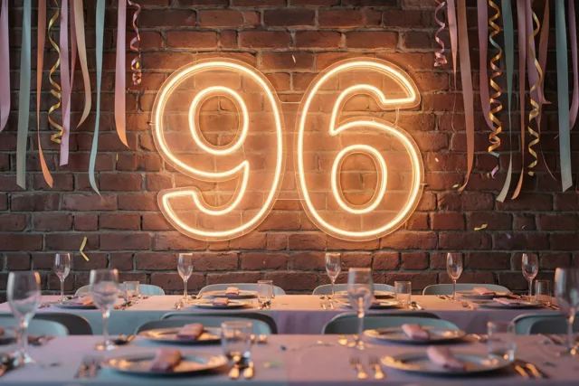 Een gloeiend neongetal 96 hangt op een bakstenen muur boven een feesttafel, met kleurrijke slingers en warme oranje, rode en zachte lavendeltinten.