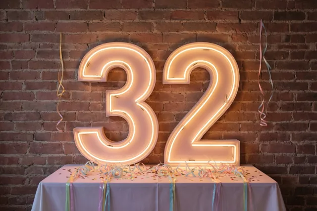 Grote neoncijfers 32 hangen aan een rode bakstenen muur boven een tafel met een wit kleed en kleurrijke slingers.