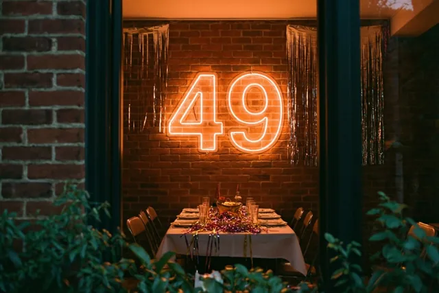 Een warm verlicht neonteken met 49 hangt aan een bakstenen muur boven een gedekte feesttafel, met donkere zuilen en groen op de voorgrond.