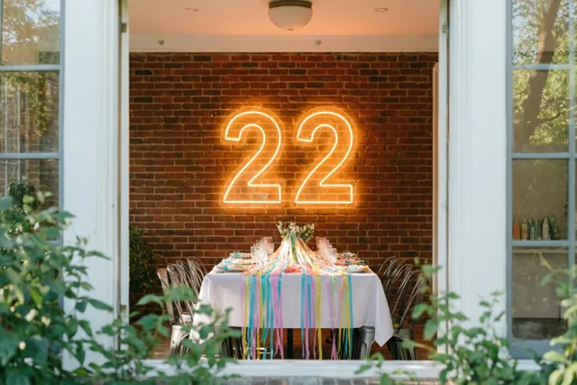 Door een open deur is een feesttafel met kleurrijke slingers te zien onder een neongetal 22 aan een bakstenen muur, met groen op de voorgrond.
