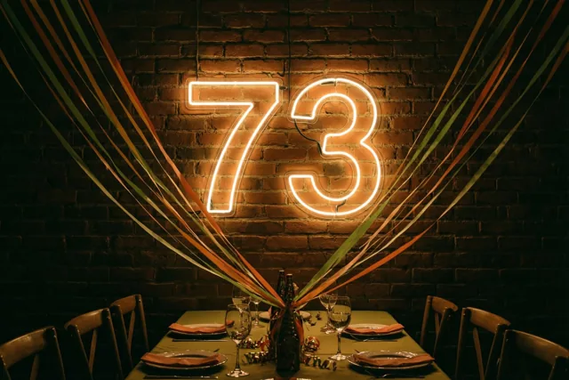 Een warm verlicht neongetal 73 hangt aan een bakstenen muur boven een gedekte feesttafel met kleurrijke slingers richting de muur.