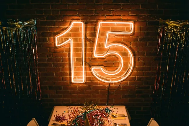 Een lichtgevend neongetal 15 hangt aan een rode bakstenen muur boven een feesttafel met kleurrijke slingers in oranje en gouden licht.