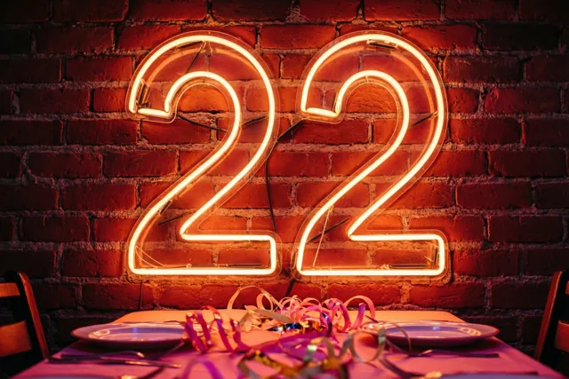 Een groot neongetal 22 hangt aan een rode bakstenen muur boven een feesttafel met borden en kleurrijke slingers.