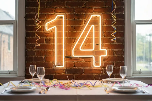 Een neongetal 14 geeft warm licht boven een tafel met borden, glazen en kleurrijke slingers tegen een rode bakstenen muur.