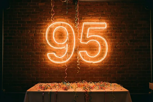Een groot neongetal 95 gloeit warm tegen een donkere bakstenen muur boven een tafel met een wit kleed en kleurrijke slingers.