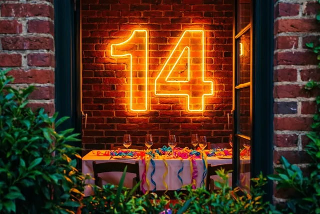 Door een donker raamkozijn is een neongetal 14 boven een feestelijk versierde tafel te zien, met een rode bakstenen muur en groen op de voorgrond.