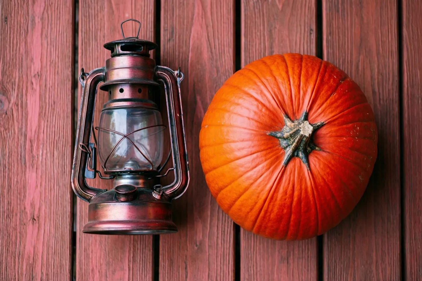 Een oude olielantaarn en een hele oranje pompoen staan naast elkaar op een rood houten oppervlak in warme herfsttinten.