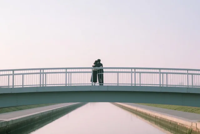 Twee mensen omhelzen elkaar in de verte op een voetbrug boven een rustig kanaal, in zachte lichtblauwe en witte tinten.