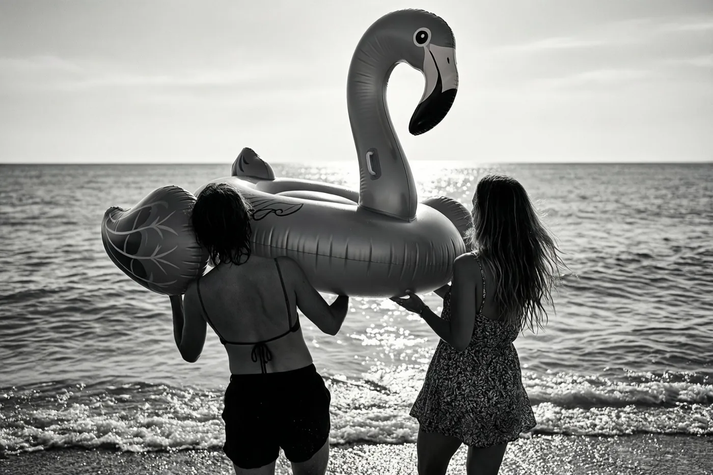 Twee mensen dragen met hun rug naar de camera een grote opblaasflamingo naar een kalme zee op een zwart-witfoto met fel zonlicht op het water.