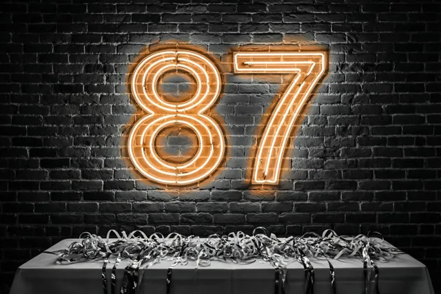 Een oranje neongetal 87 hangt aan een donkere bakstenen muur boven een tafel met zilveren slingers.