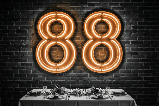 Twee grote oranje neonachten hangen aan een donkere bakstenen muur boven een formeel gedekte feesttafel in zwart-wittinten.