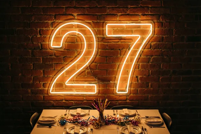 Een neonlicht in de vorm van 27 hangt aan een bakstenen muur boven een feestelijk gedekte tafel en verspreidt een warme oranje gloed.