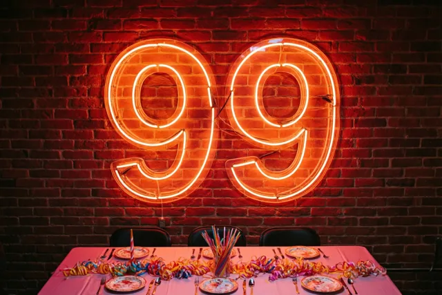 Een groot oranje neonbord met 99 hangt aan een rode bakstenen muur boven een feesttafel met borden, bestek en kleurrijke slingers in warm licht.