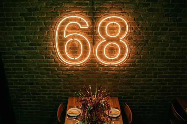 Een oranje neongetal 68 hangt op een donkere bakstenen muur boven een houten feesttafel met borden, bestek en kleurrijke slingers in het midden.