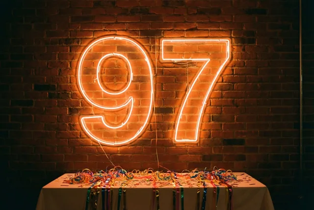Een oranje neongetal 97 hangt aan een donkere bakstenen muur boven een tafel met kleurrijke slingers.