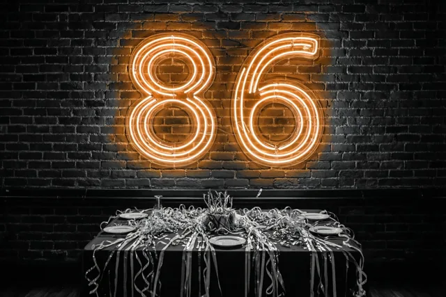 Een neon-86 in warm oranje licht hangt op een donkere bakstenen muur boven een tafel met zwart tafelkleed, zilveren slingers en borden.