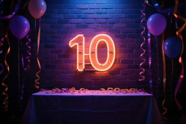 Een oranje neongetal 10 hangt aan een donkere bakstenen muur boven een tafel met een blauw kleed en slingers, tussen paarse en blauwe ballonnen.