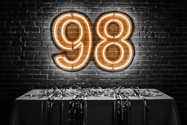 Een oranje neongetal 98 hangt aan een donkere bakstenen muur boven een tafel met een wit kleed en zilveren slingers.