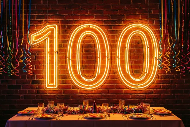 Een neongetal 100 gloeit boven een gedekte feesttafel tegen een bakstenen muur, met kleurrijke slingers aan beide kanten en warm oranje licht.