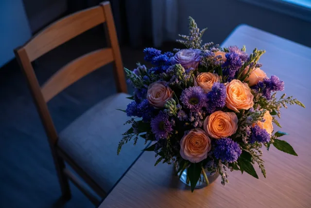 Een vaas met oranje en paarse bloemen staat op een houten tafel naast een lege houten stoel met een grijs kussen in zacht koel licht.
