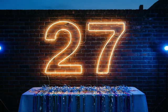 Een groot oranje neonlicht vormt 27 op een donkere bakstenen muur boven een tafel met blauwe en zilveren slingers, in blauw nachtelijk licht.