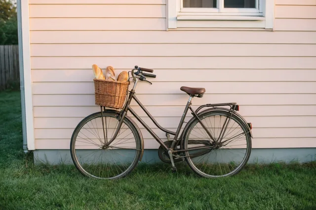 Een oude fiets met een rieten mand vol vers brood leunt tegen de lichte gevelbekleding van een huis, op een grasveld in zacht licht bij het ochtendgloren.