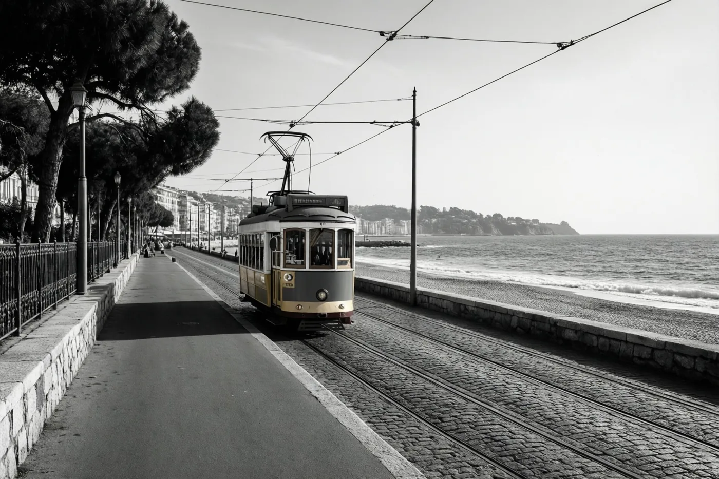 Op een zwart-witfoto rijdt een oude tram over een geplaveide promenade langs de zee.