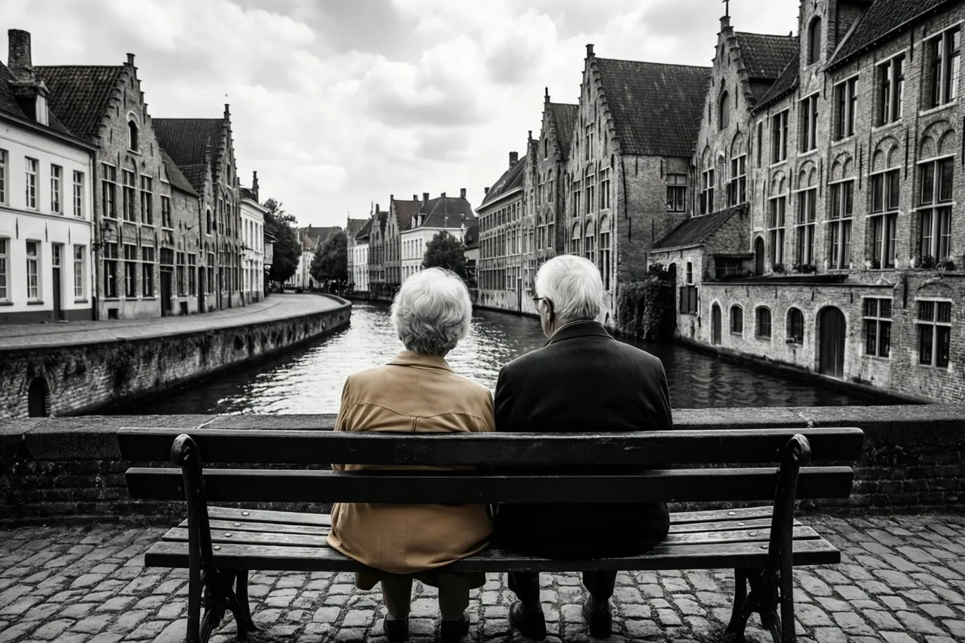 Een ouder koppel zit op een bank aan een kanaal tussen historische Vlaamse bakstenen gebouwen, in een zwart-witbeeld waarin alleen de jas van de vrouw een warme kleur heeft.