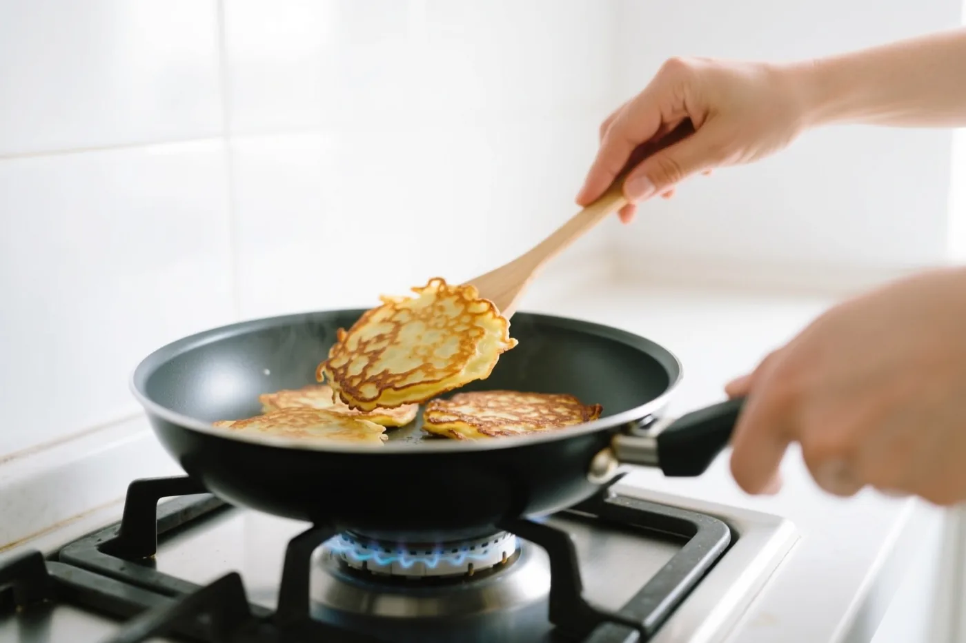 Iemand bakt goudbruine pannenkoeken in een zwarte pan op een gasvuur met een blauwe vlam in een lichte keuken met witte tegels.
