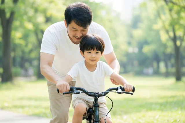 Een vader helpt zijn jonge zoon fietsen in een zonnig park vol groen.