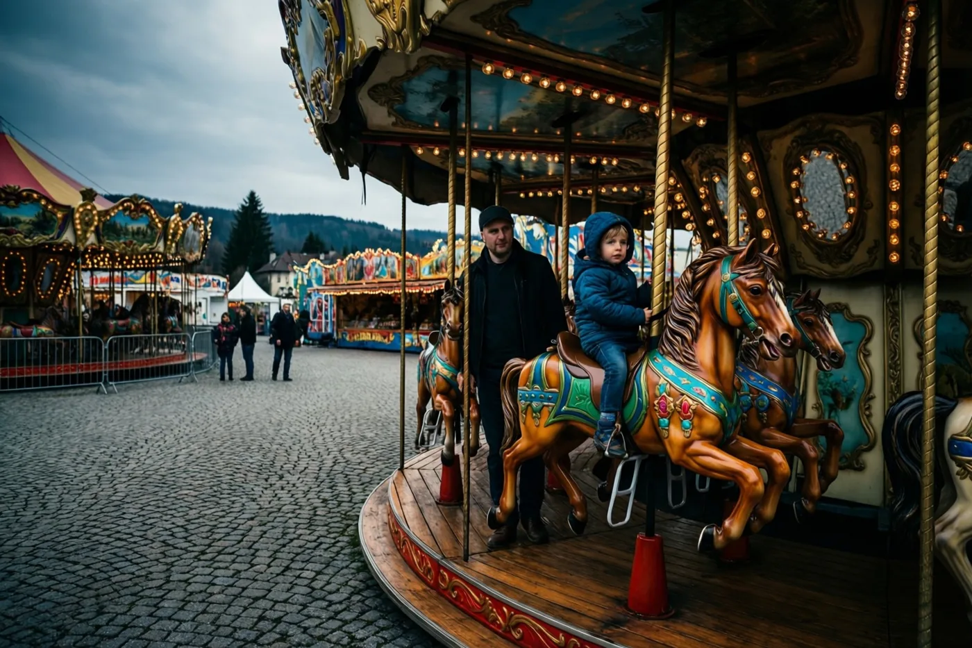 Een vader staat naast zijn kind op een draaimolenpaard op een dorpskermis met kasseien, waar de draaimolen en andere attracties warm verlicht zijn onder een bewolkte lucht.