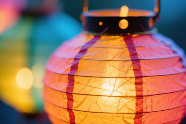 Een close-up toont een brandende papieren lantaarn met zichtbare plooien en rode ribben tegen onscherpe, kleurrijke lichtjes.