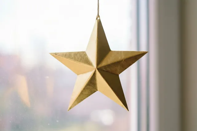 Een gouden papieren ster hangt aan een touwtje voor een helder raam in zacht, natuurlijk licht.
