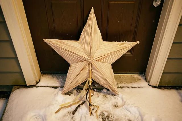 Een grote lichtgekleurde papieren ster op een stok met linten leunt tegen een donkerbruine voordeur in de sneeuw.