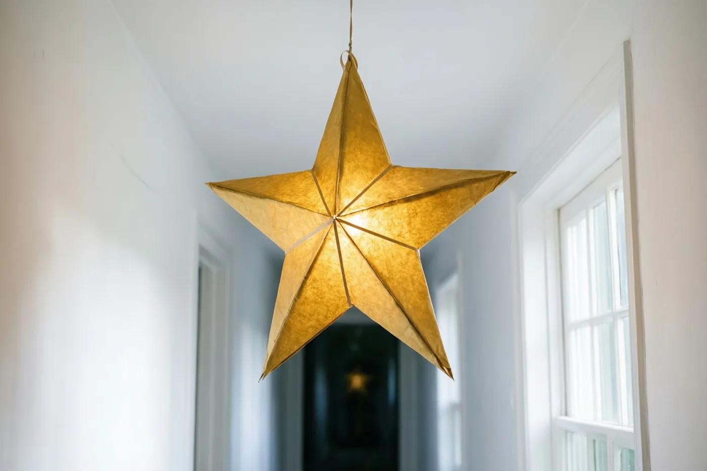 Een gouden papieren sterlantaarn hangt aan het plafond van een witte gang met zacht daglicht uit een raam aan de rechterkant.