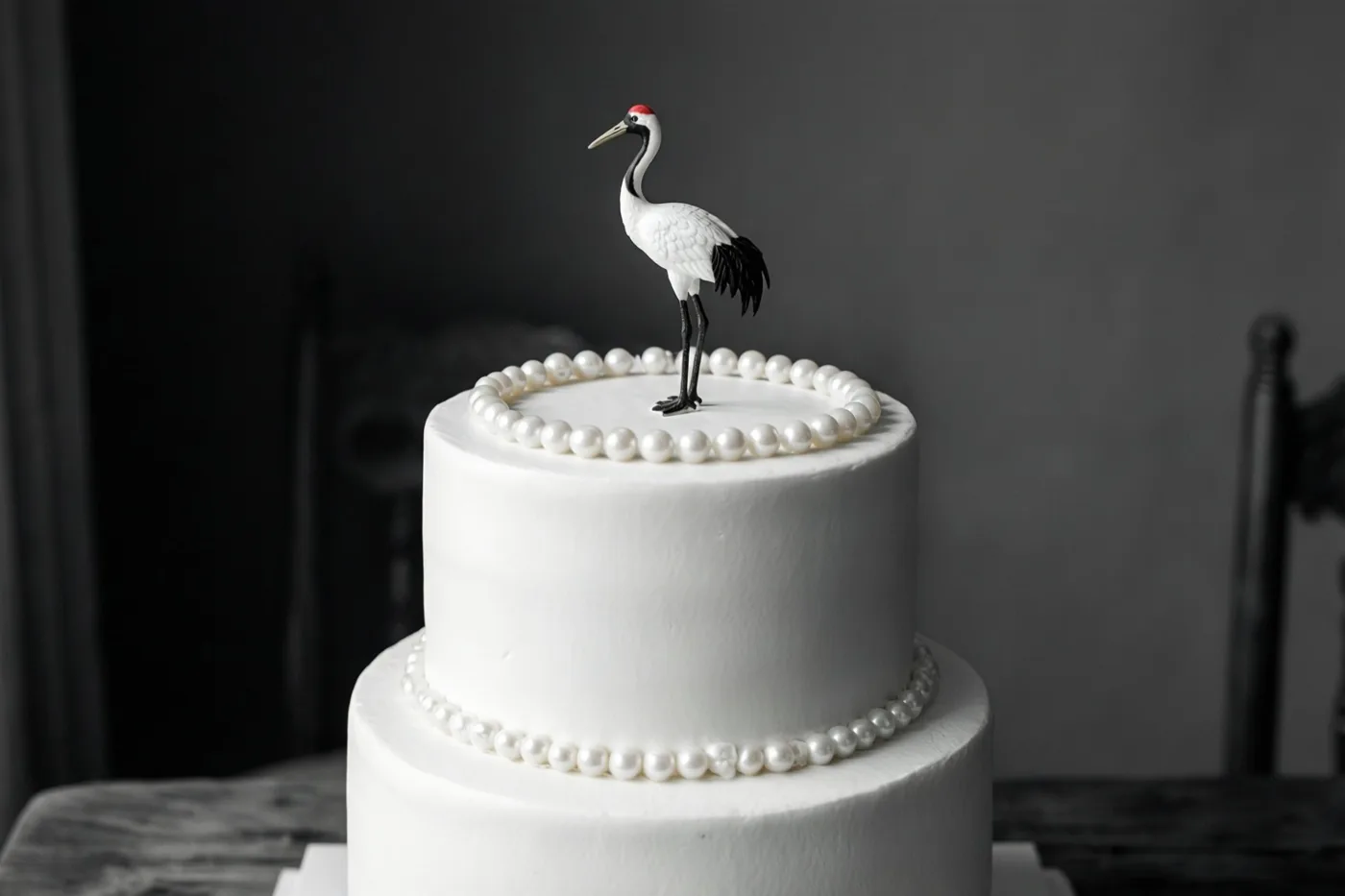 Een witte taart met twee lagen en parels rond beide lagen is bekroond met een kleine kraanvogel van suiker tegen een donkere, sobere achtergrond.
