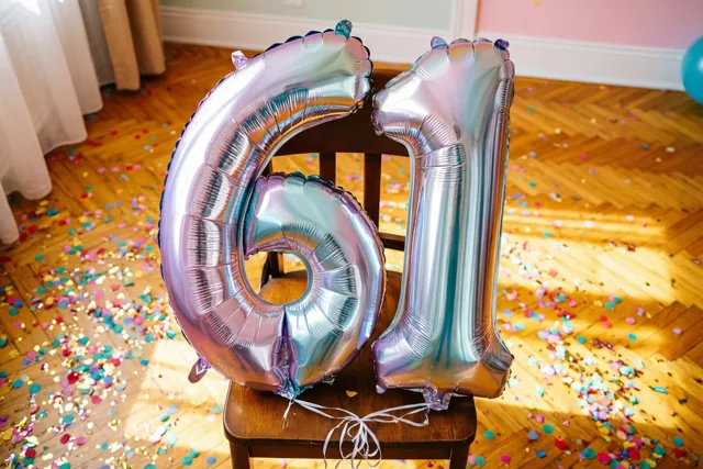 Zilveren en pastelkleurige ballonnen in de vorm van 61 zijn vastgemaakt aan een houten stoel, met gekleurde confetti op de zonverlichte houten vloer.