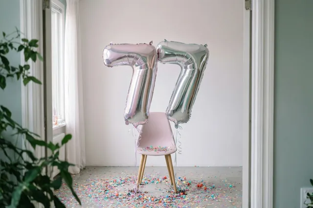 Zilveren en pastelkleurige cijferballonnen vormen 77 aan een roze stoel, met kleurrijke confetti op de vloer en daglicht door witte gordijnen.