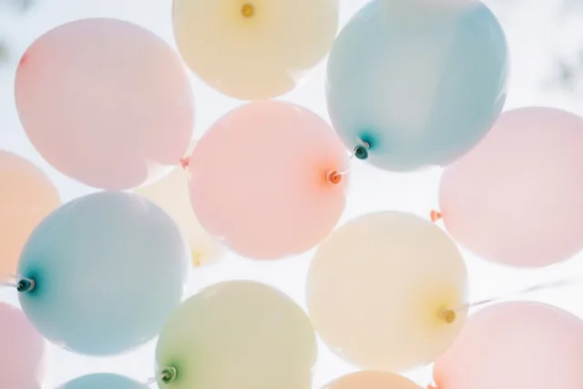 Een tros zachtroze, lichtblauwe en crèmekleurige ballonnen vult van onderaf gezien het beeld tegen een helderwitte achtergrond.
