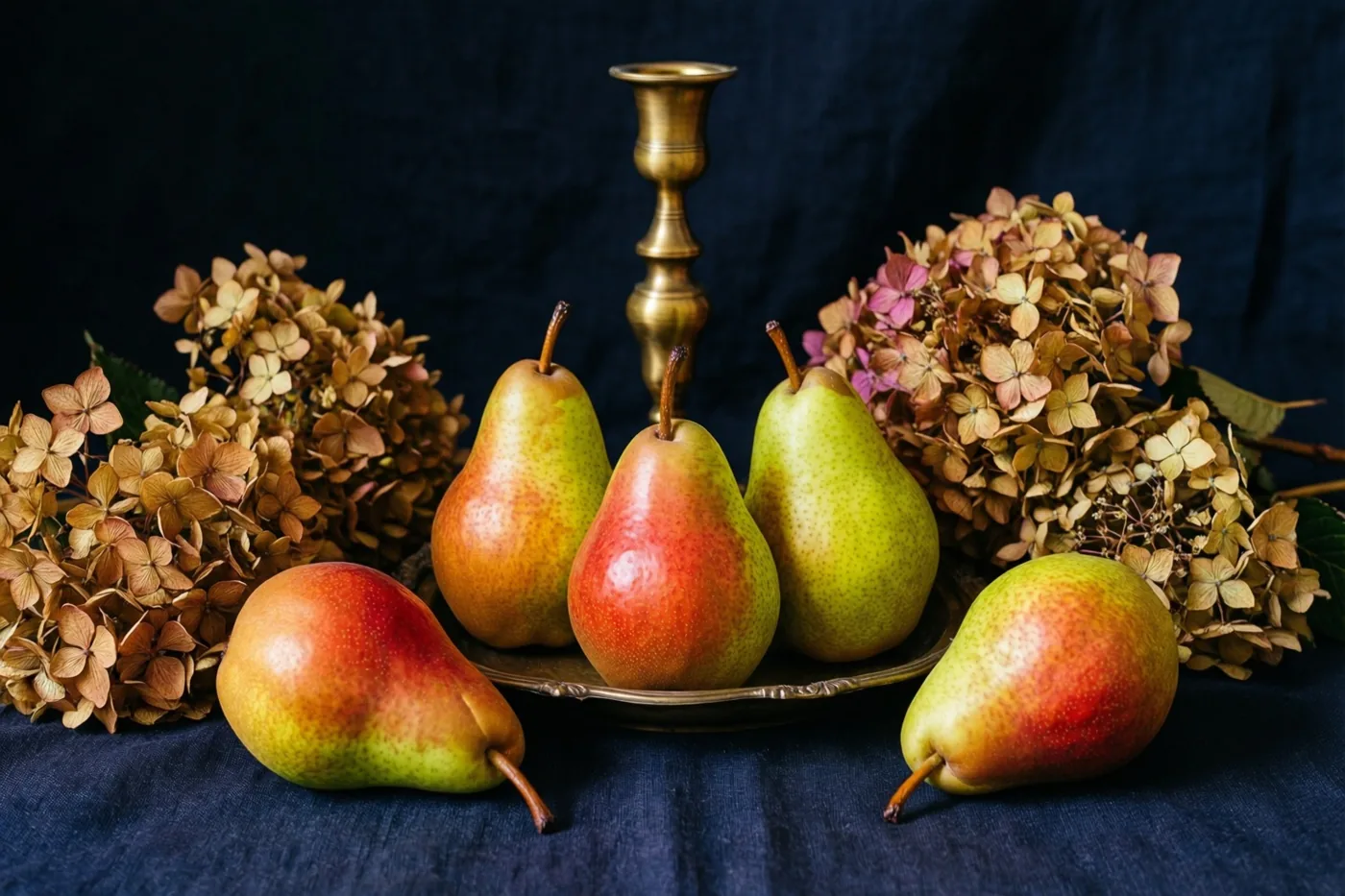 Peren, een messing kandelaar en gedroogde hortensia's liggen op donker linnen tegen een diepblauwe achtergrond, in rode, groene en gouden tinten.