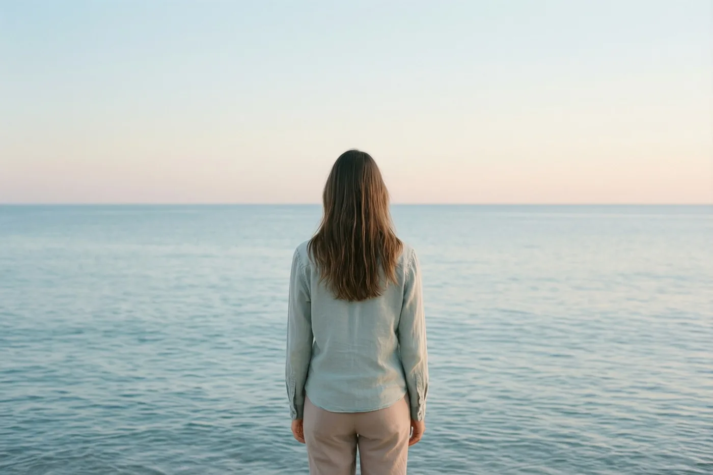 Een persoon met lang haar staat met de rug naar de kijker aan een rustige zee, in zachte blauwe en roze tinten.