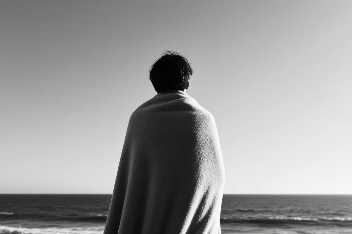 Een persoon in een deken staat met de rug naar de kijker op het strand en kijkt naar zee in een zwart-witfoto.