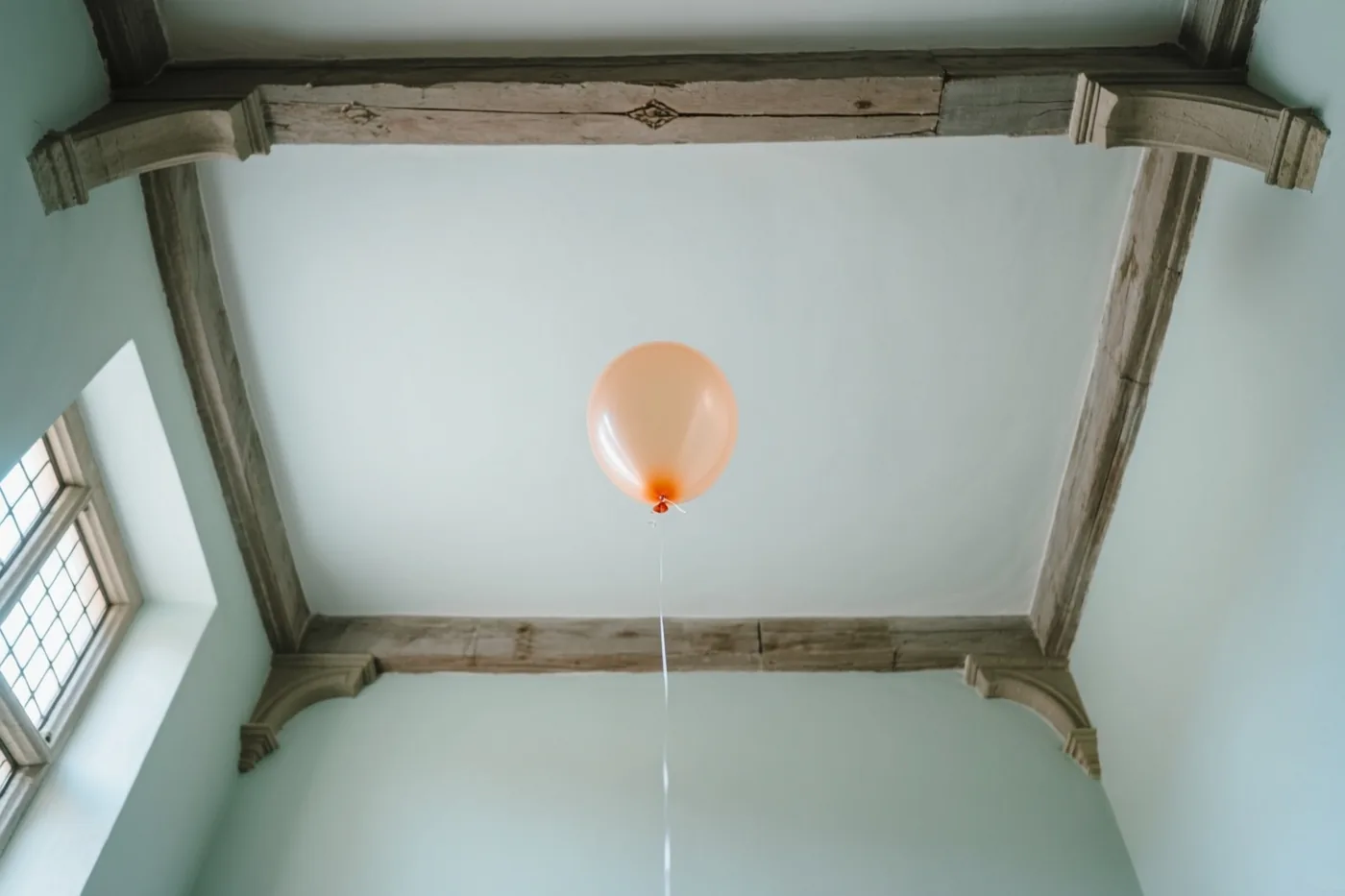Een perzikkleurige heliumballon zweeft onder een hoog plafond met verweerde houten balken in een oud Vlaams herenhuis met pastelkleurige muren en daglicht uit een raam.
